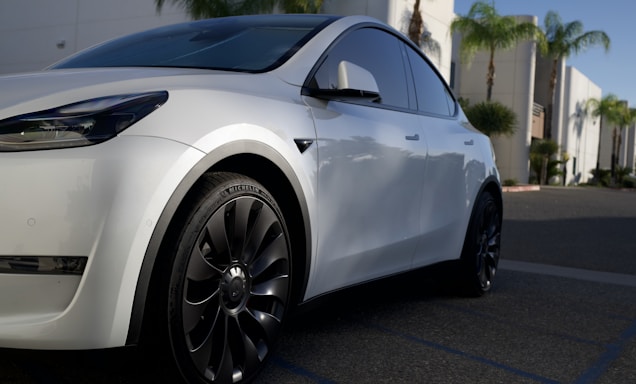 A sleek white car with modern design elements, featuring stylish black wheels. It is parked in a sunlit urban environment with palm trees visible in the background. The car's front and side are prominently displayed, highlighting its smooth curves and tinted windows.