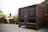 A wooden speaker cabinet with two mounted speakers placed against a background of cut tree logs. The cabinet is dark brown and appears to be positioned on a paved surface.