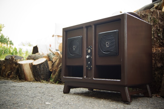 A wooden speaker cabinet with two mounted speakers placed against a background of cut tree logs. The cabinet is dark brown and appears to be positioned on a paved surface.