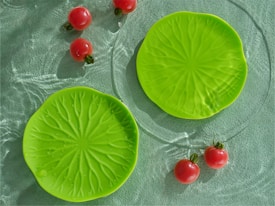 Two bright green plates designed to resemble lily pads are placed on a textured surface with subtle water ripples. Several small red tomatoes are scattered nearby, some partially submerged, enhancing a fresh and natural ambiance.