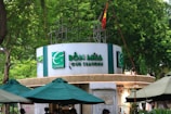 A circular kiosk is prominently displayed, featuring the signage 'Bốn Mùa - Four Seasons' in bold green letters. Surrounding the kiosk are several large green umbrellas providing shade. The background is lush with dense green foliage, and a national flag is visible atop the structure.