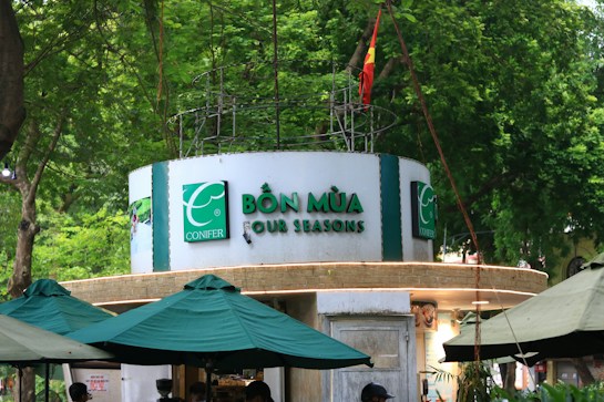 A circular kiosk is prominently displayed, featuring the signage 'Bốn M&ugrave;a - Four Seasons' in bold green letters. Surrounding the kiosk are several large green umbrellas providing shade. The background is lush with dense green foliage, and a national flag is visible atop the structure.