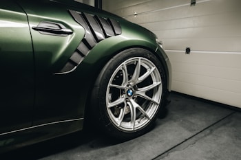 A close-up view of a luxury car's front side, highlighting the green body with carbon fiber details and a distinctive side air vent. The image prominently features a high-performance alloy wheel with a visible BMW logo at the center and sleek tire edges. The car is parked inside a garage with concrete flooring and a light-colored, paneled garage door partially visible in the background.