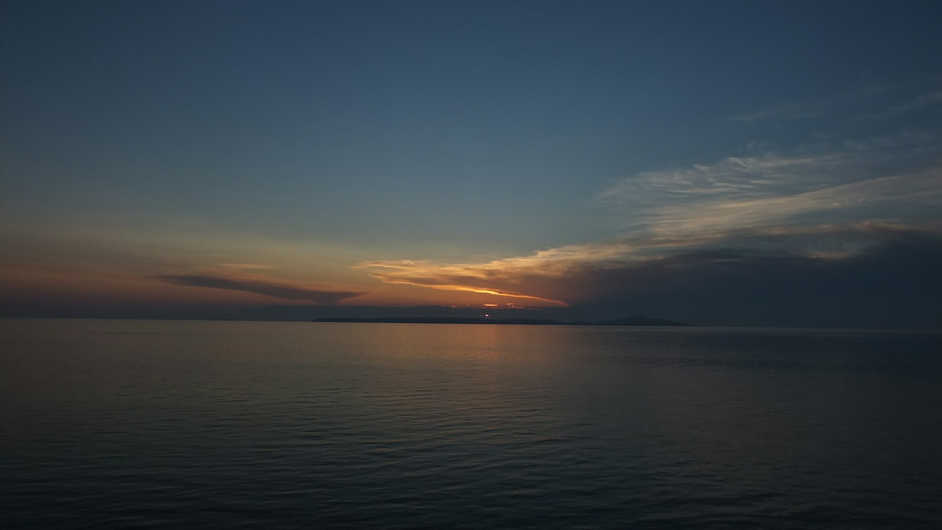 The horizon is painted with the subtle hues of a sunset, featuring a vibrant sky transitioning from deep blue to warm oranges and yellows. The sun is partially visible, casting a soft glow over the calm sea below. Wispy clouds add texture to the sky, completing the serene coastal scenery.