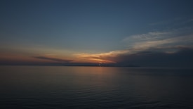 The horizon is painted with the subtle hues of a sunset, featuring a vibrant sky transitioning from deep blue to warm oranges and yellows. The sun is partially visible, casting a soft glow over the calm sea below. Wispy clouds add texture to the sky, completing the serene coastal scenery.