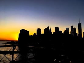 Silhouette of a city skyline against a dramatic sunset with deep orange, yellow, and blue hues filling the sky. The buildings appear in dark outlines, creating a striking contrast with the vibrant colors in the background.