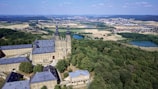 An aerial view of a historic building complex surrounded by dense woodland and open fields. The architecture features a prominent structure with two tall towers, set against a backdrop of rolling hills and a small village in the distance. Several bodies of water are scattered throughout the landscape, adding to the variety of natural elements.
