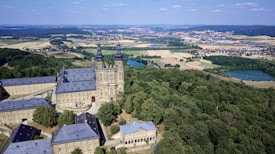 An aerial view of a historic building complex surrounded by dense woodland and open fields. The architecture features a prominent structure with two tall towers, set against a backdrop of rolling hills and a small village in the distance. Several bodies of water are scattered throughout the landscape, adding to the variety of natural elements.