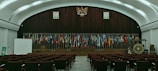 The image depicts an empty auditorium with rows of red chairs facing a stage adorned with numerous international flags. A large emblem is mounted on a wooden panel, flanked by portraits of individuals. On the stage, there is a podium, a large gong, and a screen to the left. The room features a high, arched ceiling with ambient lighting.