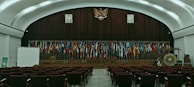 The image depicts an empty auditorium with rows of red chairs facing a stage adorned with numerous international flags. A large emblem is mounted on a wooden panel, flanked by portraits of individuals. On the stage, there is a podium, a large gong, and a screen to the left. The room features a high, arched ceiling with ambient lighting.