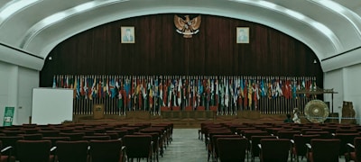 The image depicts an empty auditorium with rows of red chairs facing a stage adorned with numerous international flags. A large emblem is mounted on a wooden panel, flanked by portraits of individuals. On the stage, there is a podium, a large gong, and a screen to the left. The room features a high, arched ceiling with ambient lighting.