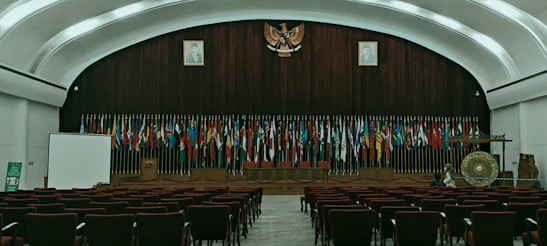 The image depicts an empty auditorium with rows of red chairs facing a stage adorned with numerous international flags. A large emblem is mounted on a wooden panel, flanked by portraits of individuals. On the stage, there is a podium, a large gong, and a screen to the left. The room features a high, arched ceiling with ambient lighting.