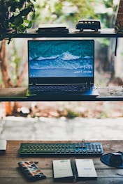 A cozy wooden desk with an open laptop, stacked books, and a large window showing a serene beach view.