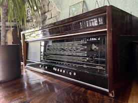 An antique wooden radio sits on a wooden surface, featuring a black front panel with dials and vintage tuning indicators. The surrounding decor includes a potted plant with long, thin leaves and a textured stone wall with subtle greenish tones in the background.