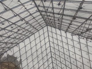 A glass and metal structural design with a triangular pattern, intersecting lines, and a cloudy sky visible through the transparent panes. In the lower corner, part of a historic building with ornate architecture is visible.
