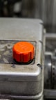A close-up image of a metallic surface featuring an orange oil cap or plug. The cap appears to be hexagonal, contrasting with the weathered and industrial surface it is mounted on.