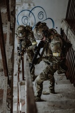 Two soldiers in camouflage gear are ascending a concrete staircase inside a worn and graffiti-decorated building. They are armed, wearing helmets, and appear to be in a tactical operation.