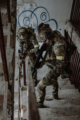 Two soldiers in camouflage gear are ascending a concrete staircase inside a worn and graffiti-decorated building. They are armed, wearing helmets, and appear to be in a tactical operation.