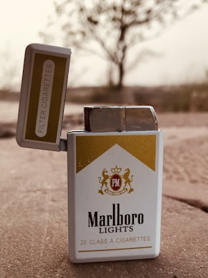 An open pack of Marlboro Lights cigarettes is positioned on a stone surface with a blurred natural setting in the background. The pack design features a gold and white color scheme with branding and text visible.