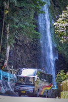 A large colorful bus is driving past a scenic waterfall. The waterfall cascades down a rocky cliff surrounded by lush greenery. Tall trees and vibrant foliage frame the scene, creating a natural backdrop.
