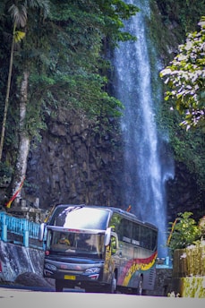 A large colorful bus is driving past a scenic waterfall. The waterfall cascades down a rocky cliff surrounded by lush greenery. Tall trees and vibrant foliage frame the scene, creating a natural backdrop.