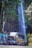 A large colorful bus is driving past a scenic waterfall. The waterfall cascades down a rocky cliff surrounded by lush greenery. Tall trees and vibrant foliage frame the scene, creating a natural backdrop.
