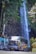 A large colorful bus is driving past a scenic waterfall. The waterfall cascades down a rocky cliff surrounded by lush greenery. Tall trees and vibrant foliage frame the scene, creating a natural backdrop.