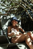 A woman is lounging on a sunbed in a shaded outdoor area, wearing a dark sunhat and a bikini. She is holding a stuffed animal against her belly, suggesting a calm and relaxed atmosphere with patterned shadows from overhead trees falling on her skin.