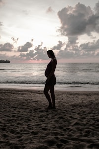 A serene coastal landscape at sunset with a pregnant woman posing gently by the shore.
