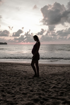 Pregnant woman walking barefoot along a sandy beach with waves gently touching her feet under a cloudy sky.
