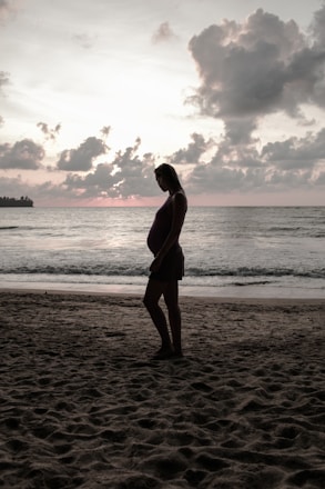 A pregnant woman stands on a sandy beach with the ocean in the background. The sky is partly cloudy and the sun appears to be setting, creating a calm and serene atmosphere.