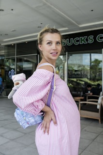 A pregnant woman with blonde hair is wearing a pink and white striped dress and holding a child's toy and a small, colorful bag. She is standing outside a coffee shop with a seating area and some greenery visible through large windows in the background.