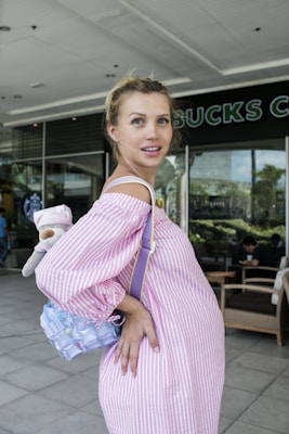 A pregnant woman with blonde hair is wearing a pink and white striped dress and holding a child's toy and a small, colorful bag. She is standing outside a coffee shop with a seating area and some greenery visible through large windows in the background.