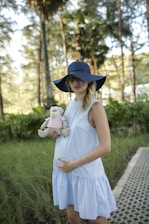 A pregnant woman wearing a light blue dress and a wide-brimmed blue hat stands in a natural setting with trees in the background. She holds a stuffed toy dressed in a pink outfit, and her hand gently rests on her belly. The setting conveys a serene and peaceful atmosphere.