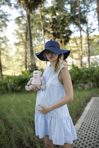 A pregnant woman wearing a light blue dress and a wide-brimmed blue hat stands in a natural setting with trees in the background. She holds a stuffed toy dressed in a pink outfit, and her hand gently rests on her belly. The setting conveys a serene and peaceful atmosphere.