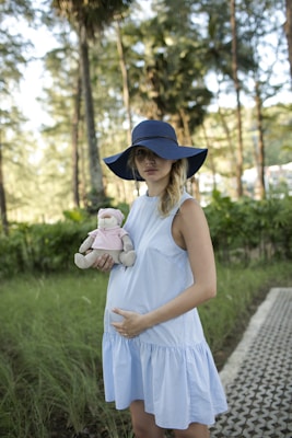 A pregnant woman wearing a light blue dress and a wide-brimmed blue hat stands in a natural setting with trees in the background. She holds a stuffed toy dressed in a pink outfit, and her hand gently rests on her belly. The setting conveys a serene and peaceful atmosphere.