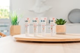 Four tubes of skincare product labeled 'BB Crème' stand on a round wooden tray. In the background, there are green plants in decorative pots. The setting appears to be a bright, minimalist indoor environment with soft lighting.