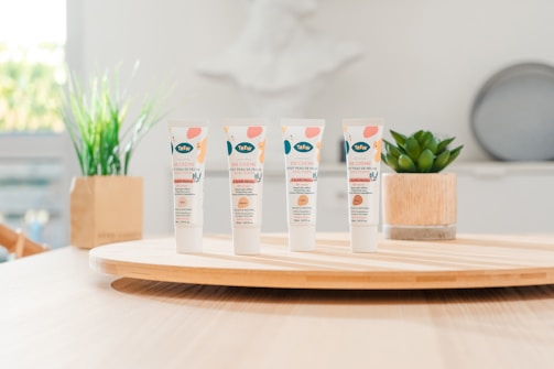 Four tubes of skincare product labeled 'BB Crème' stand on a round wooden tray. In the background, there are green plants in decorative pots. The setting appears to be a bright, minimalist indoor environment with soft lighting.