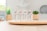 Four tubes of skincare product labeled 'BB Crème' stand on a round wooden tray. In the background, there are green plants in decorative pots. The setting appears to be a bright, minimalist indoor environment with soft lighting.