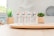 Four tubes of skincare product labeled 'BB Crème' stand on a round wooden tray. In the background, there are green plants in decorative pots. The setting appears to be a bright, minimalist indoor environment with soft lighting.