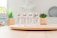 Four tubes of skincare product labeled 'BB Crème' stand on a round wooden tray. In the background, there are green plants in decorative pots. The setting appears to be a bright, minimalist indoor environment with soft lighting.