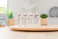 Four tubes of skincare product labeled 'BB Cr&egrave;me' stand on a round wooden tray. In the background, there are green plants in decorative pots. The setting appears to be a bright, minimalist indoor environment with soft lighting.