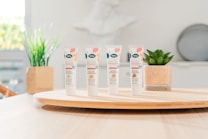 Four tubes of skincare product labeled 'BB Cr&egrave;me' stand on a round wooden tray. In the background, there are green plants in decorative pots. The setting appears to be a bright, minimalist indoor environment with soft lighting.