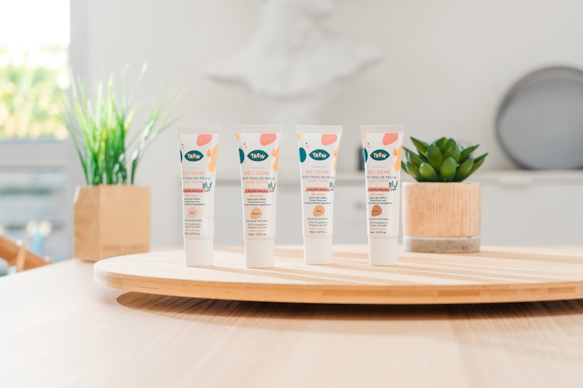 Four tubes of skincare product labeled 'BB Cr&egrave;me' stand on a round wooden tray. In the background, there are green plants in decorative pots. The setting appears to be a bright, minimalist indoor environment with soft lighting.