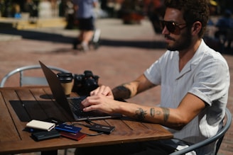 A person with tattoos is sitting at an outdoor cafe, typing on a laptop. The person is wearing sunglasses and a light short-sleeved shirt. On the table are several external hard drives and a camera, along with a coffee cup. The setting appears relaxed and sunny.