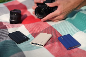 A hand holding a camera is poised above a plaid blanket. There are three Samsung portable SSDs placed on the blanket in black, white, and blue colors. A camera lens lies nearby, slightly out of focus.