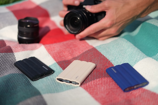 A hand holding a camera is poised above a plaid blanket. There are three Samsung portable SSDs placed on the blanket in black, white, and blue colors. A camera lens lies nearby, slightly out of focus.