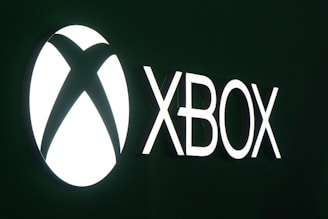 The image features a bright white Xbox logo on a dark green background. The word 'XBOX' is prominently displayed in bold letters next to the iconic circular logo.