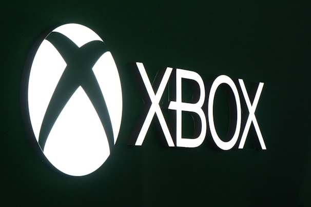 The image features a bright white Xbox logo on a dark green background. The word 'XBOX' is prominently displayed in bold letters next to the iconic circular logo.