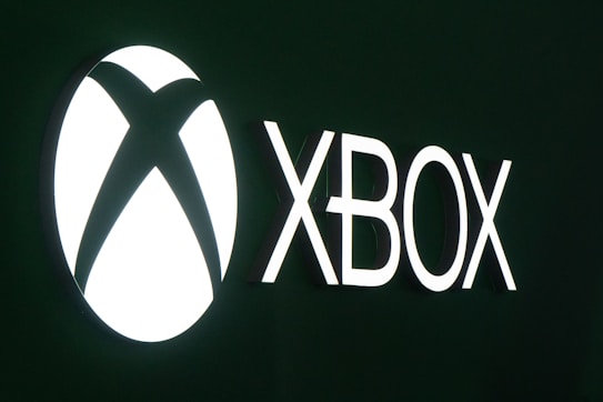 The image features a bright white Xbox logo on a dark green background. The word 'XBOX' is prominently displayed in bold letters next to the iconic circular logo.